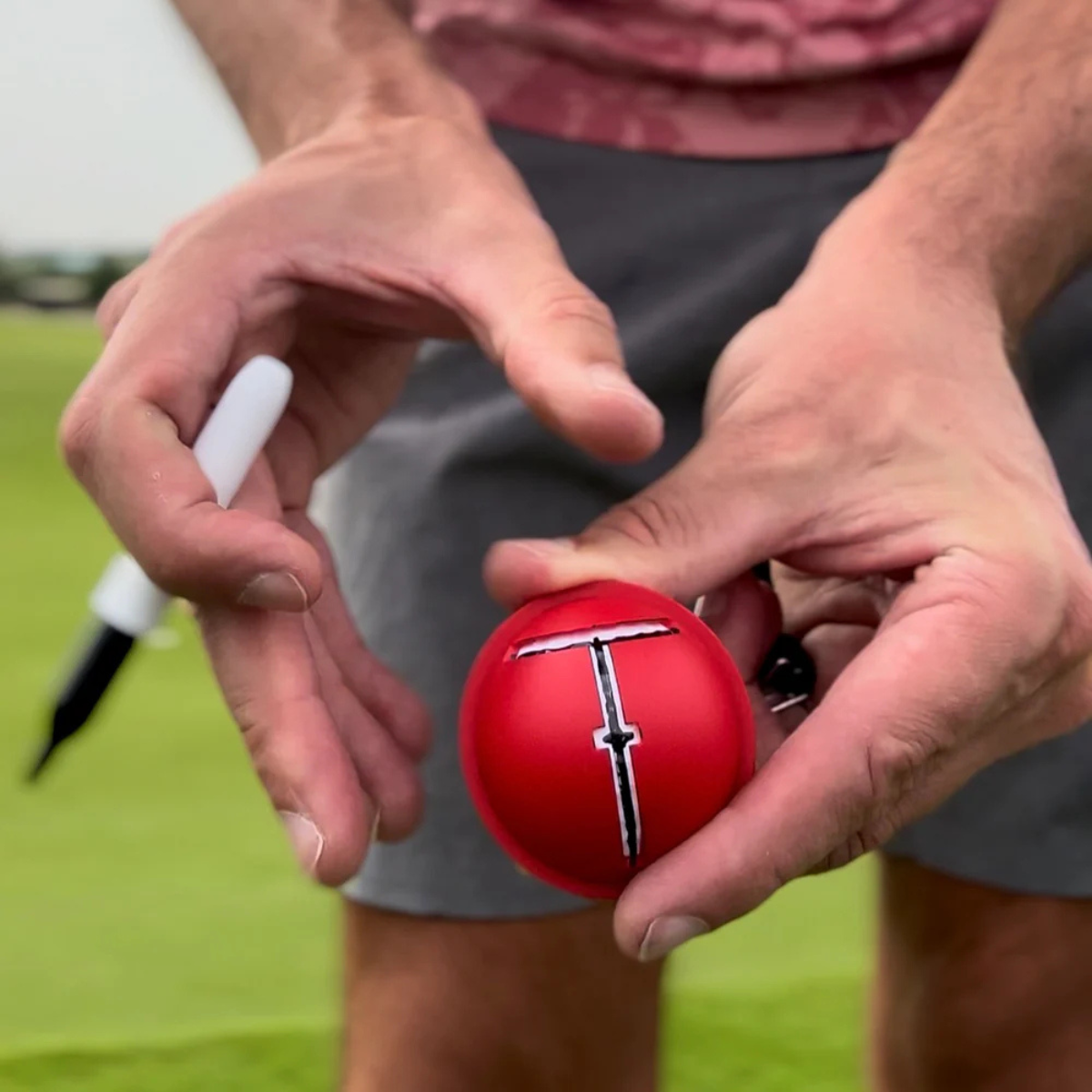Person holding a golf ball with a red cover and with a white marker on a grassy field