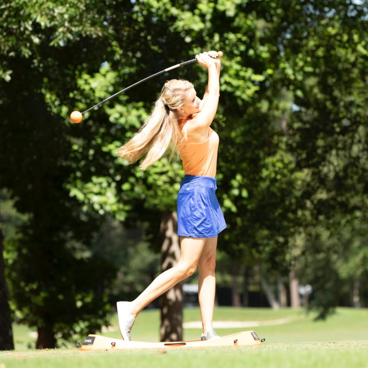 Woman playing golf with the Orange Whip on a sunny day with trees in the background