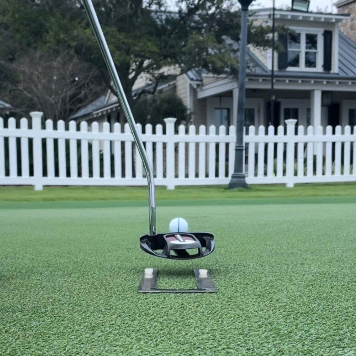 Golf putter and ball on a green putting surface with a white fence and house in the background.