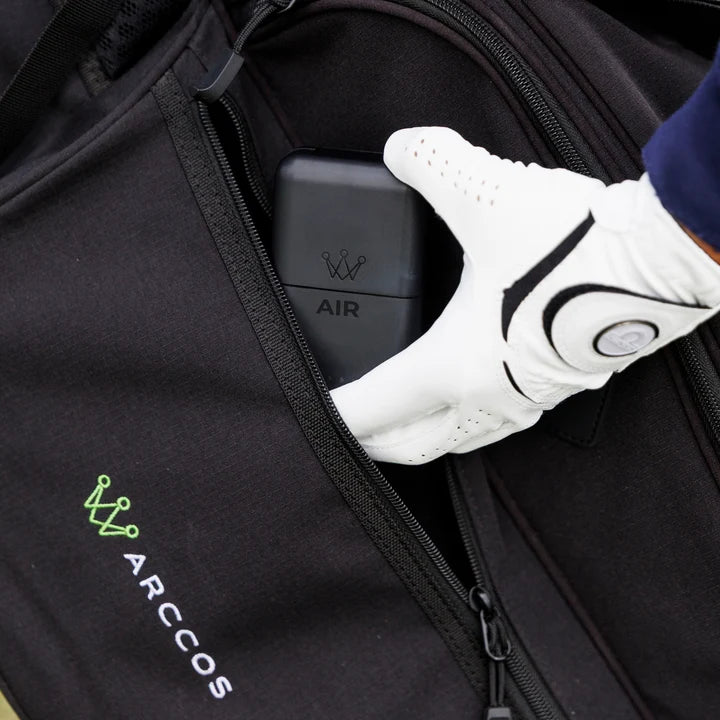 A close-up of a black Arccos golf bag showing a zippered pocket with a white golf glove holding the charging case for the Arccos Air