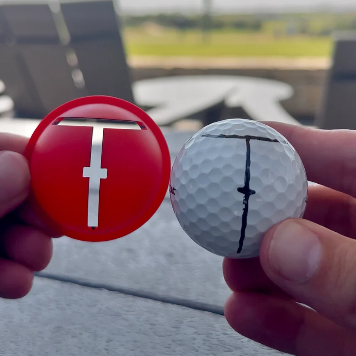 Red golf ball marker with a white golf ball held by hands outdoors.