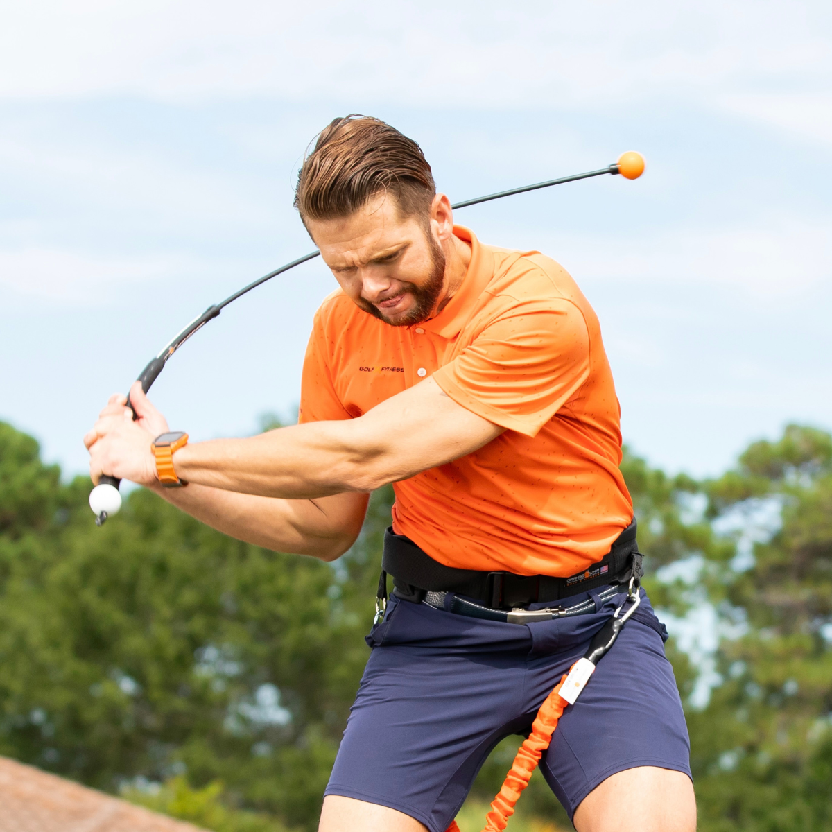 Man in orange shirt and blue shorts swinging a golf club with trees in the background