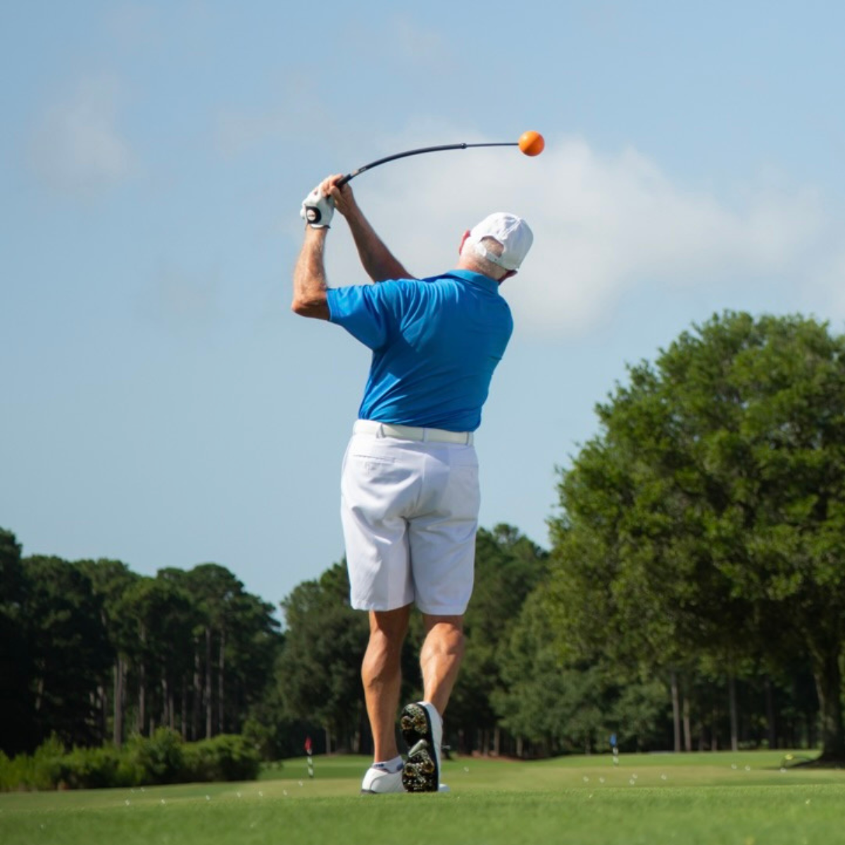 Golfer in blue shirt and white shorts swinging a golf club on a green course with trees in the background.