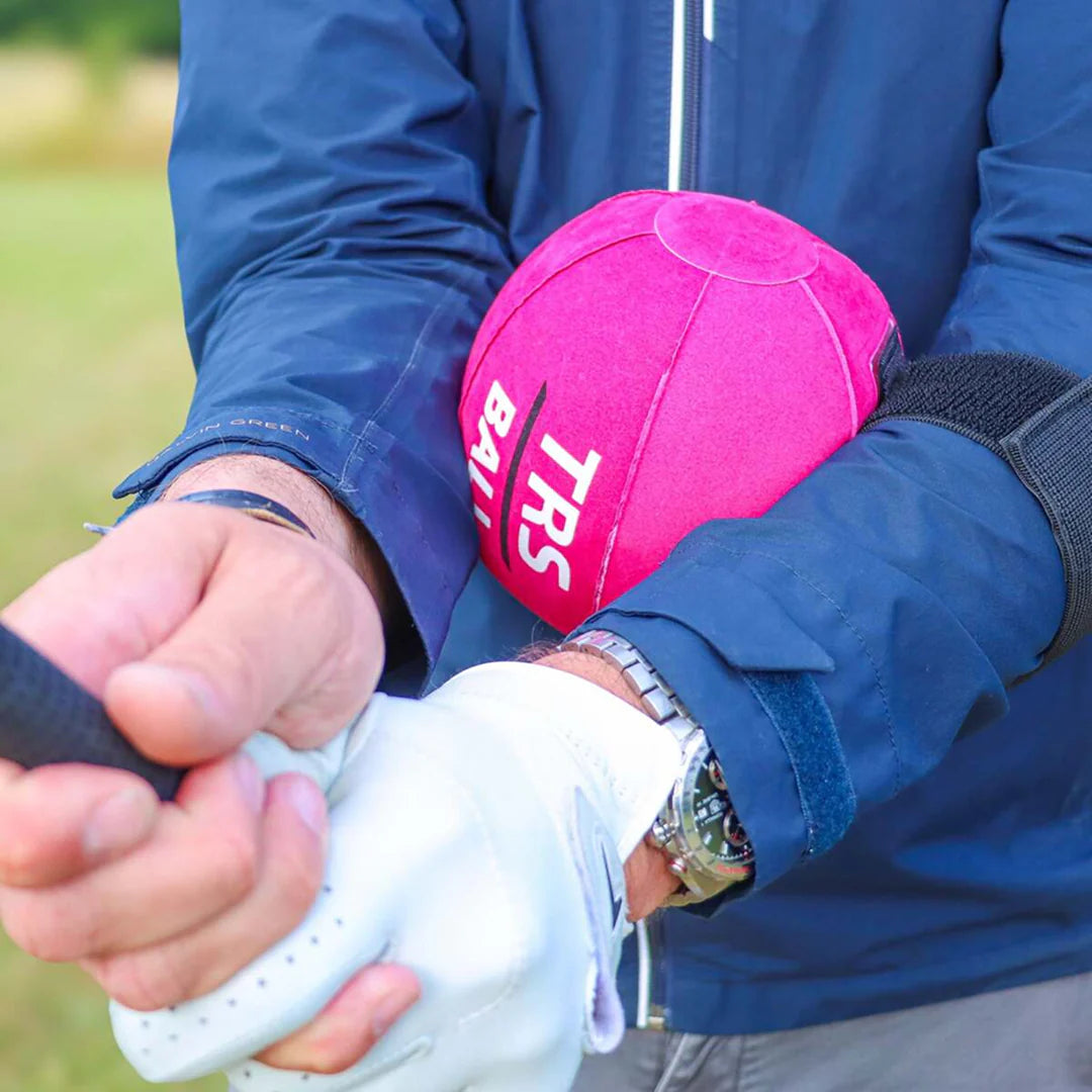 A close-up lifestyle shot of a person on a golf course holding a bright pink golf ball cap with white 'TRS BALL' branding next to a white golf glove.