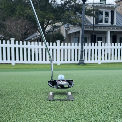 Golf putter and ball on a green putting surface with a white fence and house in the background.